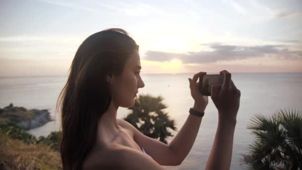 女性手拿着智能手机，做自然摄影。年轻的女游客在海滩上用手机摄像，拍下了令人惊奇的日落. — 图库视频影像