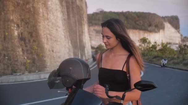 Attraente ragazza con i capelli castani si siede su una moto mette su un casco nero dello sfondo stradale. 4k — Video Stock
