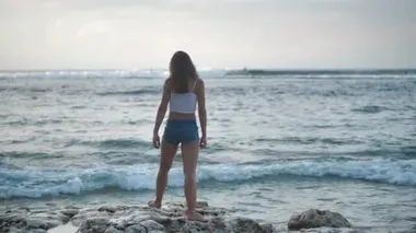 Taş plajdaki kot şortlu kız. Dalgalar kıyıya çarpıyor. 4k