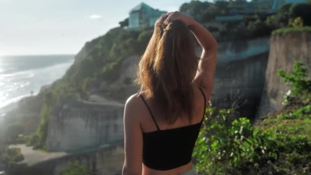 Portrait d'une fille qui se tient au-dessus d'une falaise et regarde l'océan. Ses cheveux soufflent le vent. Coucher de soleil fond 4k — Video