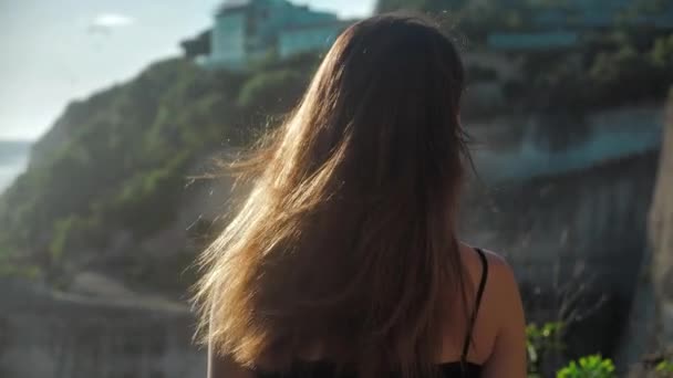 Крупным планом портрет девушки, которая стоит над скалой и смотрит на океан. Её волосы как ветер. Закат 4k — стоковое видео