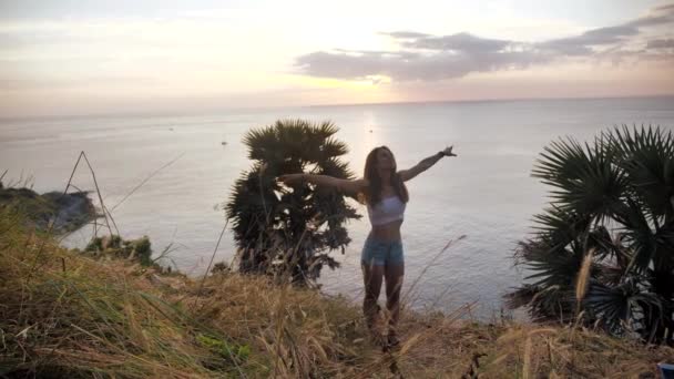 Портрет дівчини, яка стоїть над скелею і дивиться на океан. Її волосся віє. Захід сонця 4k — стокове відео