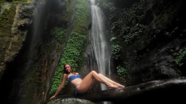 A mulher atraente senta-se em um log pelo lago em uma floresta tropical em um fundo de cachoeira na chuva. Unidade com a natureza 4k — Vídeo de Stock