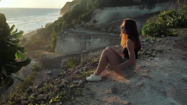 Νεαρή γυναίκα κάθεται σε ένα φόντο βράχου και απολαμβάνει την ομορφιά της φύσης. Η γυναίκα κοιτάζει τον ωκεανό. Φόντο ηλιοβασιλέματος. — Αρχείο Βίντεο