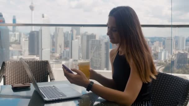 Mujer atractiva se sienta en un café con una computadora portátil, hace clic en un teléfono inteligente del fondo del rascacielos 4k — Vídeo de stock