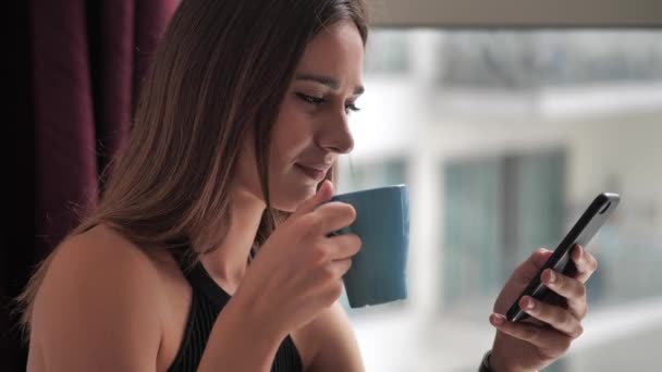 Девушка пьет кофе со смартфоном на фоне панорамного окна 4k — стоковое видео