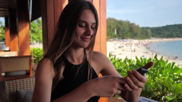 Closeup of smiling young elegant attractive girl with deep brown eyes in black dress trying to buy something enjoyable with her smartphone ocean background. Slow motion 4k — Stock Video