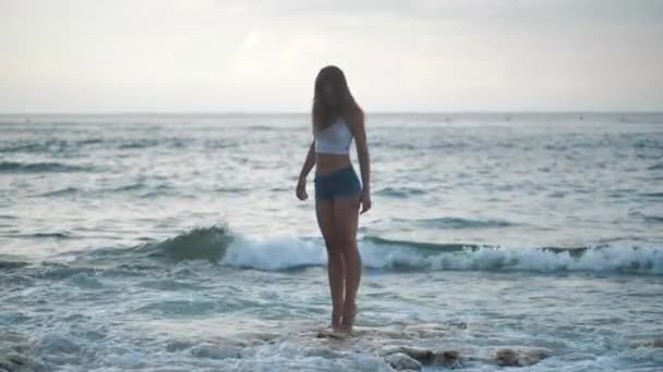 Ragazza a piedi per pantaloncini di jeans mare sulla spiaggia di pietra. Onde che si schiantano contro la riva. 4k — Video Stock