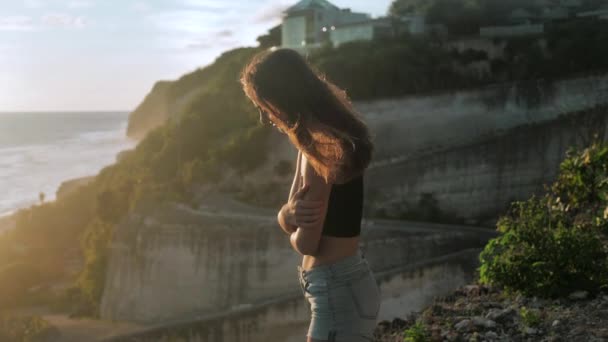 Portrait d'une fille qui se tient au-dessus d'une falaise et regarde l'océan. Ses cheveux soufflent le vent. Coucher de soleil fond 4k — Video