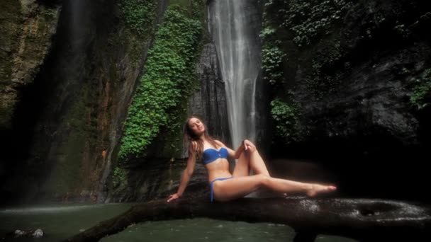 Belle femme assise en maillot de bain au bord du lac dans une forêt tropicale sur un fond de cascade sous la pluie. Unité avec la nature 4k — Video
