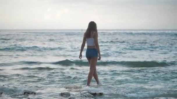 Chica en caminar a pantalones cortos de mezclilla mar en la playa de piedra. Olas chocando contra la orilla. 4k — Vídeo de stock