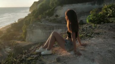 Genç kız kayanın arka planında oturuyor ve doğanın güzelliğinin tadını çıkarıyor. Kadın okyanusa bakıyor. Günbatımı arka planı. 4k