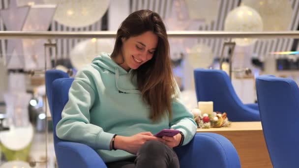 Porträt einer jungen Frau in einem türkisfarbenen Sweatshirt. Sie sitzt in einem Duty Free und klickt auf ein Smartphone. Sie erwartet ihre Flucht — Stockvideo