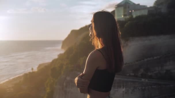 Portré egy lányról, aki egy szikla felett áll, és az óceánt nézi egy sziklának támasztva. A haja a szélben fejlődik. Naplemente háttér 4k — Stock videók