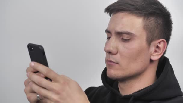 Μια σοβαρή νεαρή φορώντας μαύρο φούτερ χρησιμοποιεί ένα smartphone απομονωμένο πάνω από λευκό φόντο στον τοίχο 4k — Αρχείο Βίντεο