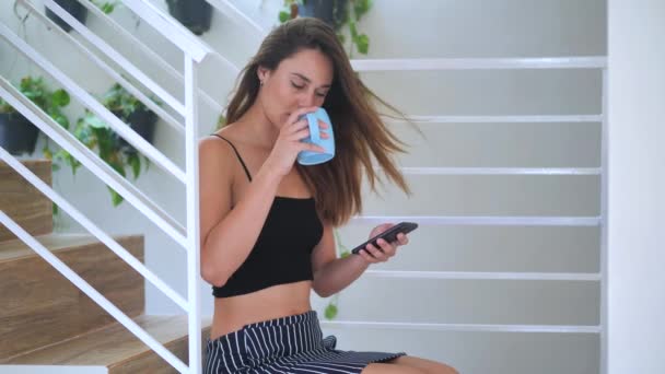 Young girl sitting on the steps clicks smartphone and drinks coffee 4k — Stock Video