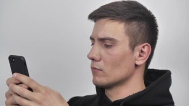 Siyah kazak giyen genç bir delikanlı beyaz duvar arkasında izole edilmiş bir akıllı telefon kullanıyor.