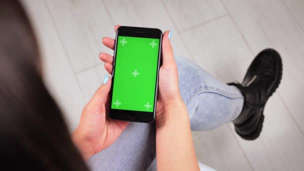 Tânăra stă acasă pe scaun și folosește un smartphone cu ecran verde. Mockup verde pentru a insera aplicații sau imagini. 4k — Videoclip de stoc