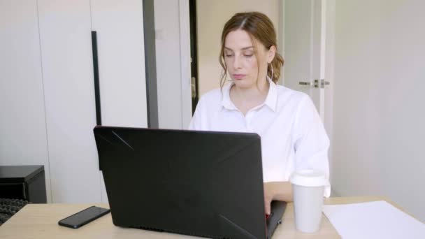 Mujer joven y seria está trabajando con el ordenador portátil en la oficina sentado en el escritorio en la habitación contemporánea. Concepto de trabajo. 4k — Vídeos de Stock