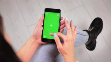 Genç kız evde sandalyeye oturup yeşil ekranlı akıllı telefon kullanıyor. Uygulamalar ya da resimler eklemek için yeşil model. 4k