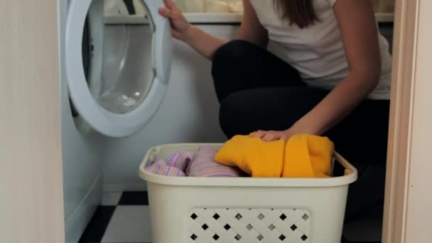 美丽的年轻女子坐在洗衣机旁边的膝盖上。她用肮脏的衣服把洗碗机装上了 — 图库视频影像