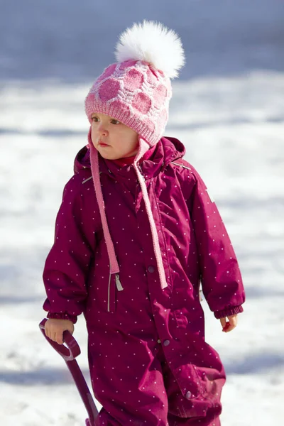 在寒冷阳光明媚的冬日 穿着鲜艳的雪衣 头戴粉色帽子 头戴白色大花冠 拿铲子玩耍的小女孩很可爱 — 图库照片