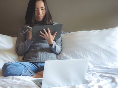 Asyalı, sıradan giyinen güzel bir kadın bluetooth kalemle tablet kullanıyor. Yatakta fotokopi odası olan bir odada. Yumuşak odak.
