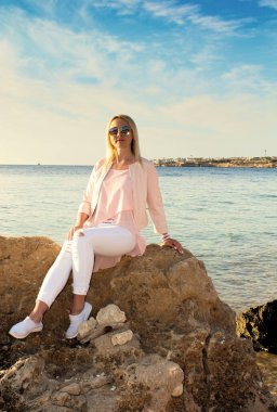 Güneş gözlüklü sarışın kız denizin yakınındaki bir taşın üzerinde oturuyor..