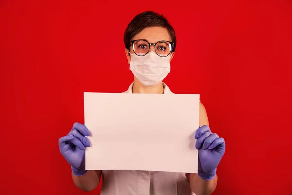 Una enfermera con una máscara protectora médica sostiene una sábana blanca en blanco sobre un fondo rojo . —  Fotos de Stock