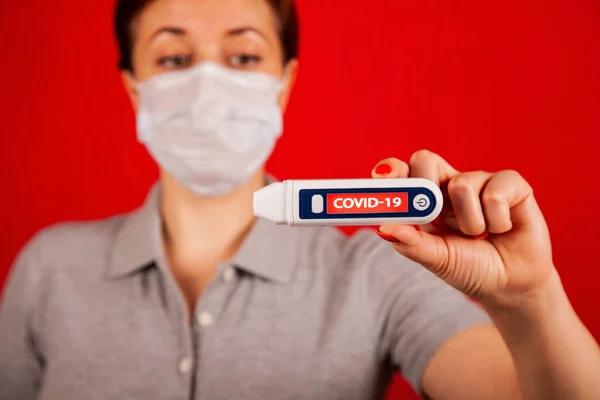 Eine Frau in Panik vor einer Infektion mit dem Coronavirus COVID-19. Eine Frau in Maske auf rotem Grund mit einem Coronavirus liest ihre Temperatur auf einem Thermometer mit der Aufschrift COVID-19 ab. — Stockfoto