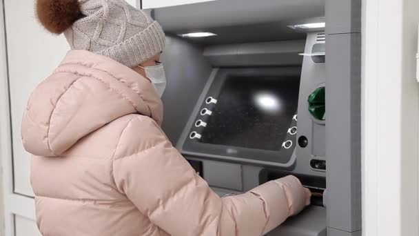 Kobieta w masce ochronnej wypłaca pieniądze z bankomatu. Wypłaty gotówki w czasie epidemii pandemii zatoczki-19 — Wideo stockowe