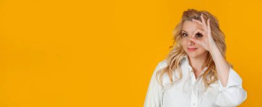 Sarışın kadın iyimser bir şekilde mutlu. Gözlerinin üzerinde bir çember çiziyor ve mutlu bir şekilde gülümsüyor. İyimser bir görünüm sergiliyor.