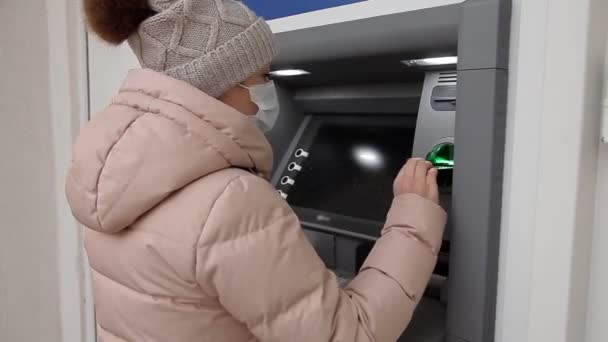 En kvinna i skyddsmask tar ut pengar från en bankomat. Kontantuttag under den feber19-pandemiska epidemin — Stockvideo