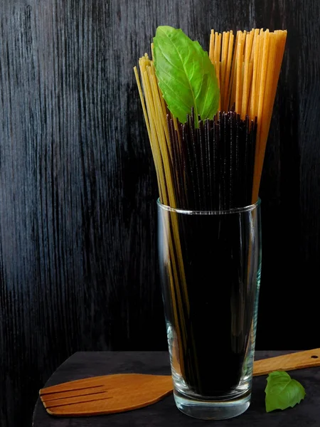 Different sorts of spaghetti in a glass decorated with basil leaves on a dark wooden background