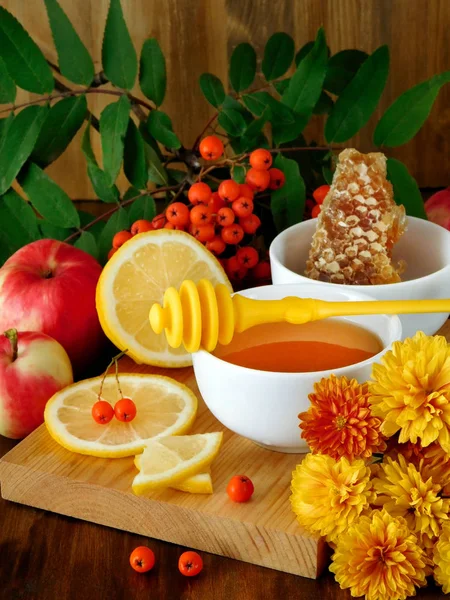 Liquid honey, lemon, apples and rowan berries surrounded by flowers on a wooden board
