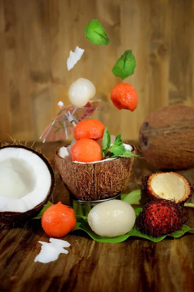 Tropical fruits: longan, rambutan and papaya ball-shaped pieces are falling into the coconut cup and around it. Ingredients for a cocktail or a dessert