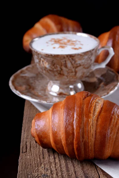 Golden croissants of yeast dough and a cup of coffee on the edge of the wooden table against black background