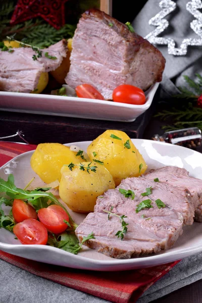 Roasted pork meat cut into slices served with potato, tomato and arugula on the white ceramic plate. Festive meal