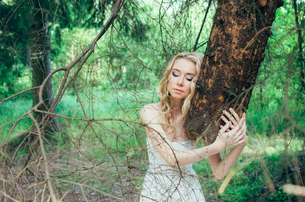 Portrait of young blond bride in forest holding tree trunk — Stock Photo, Image