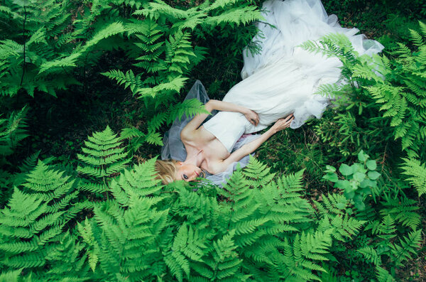 Portrait of young blond bride that lies on earth in forest among fern, looking to the camera