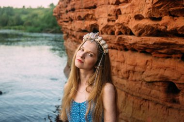 Mermilerden taç giymiş genç bir kadın kırmızı kum duvarın yanında duruyor.