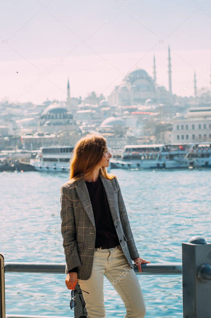 Beautiful girl with blond hair stands on embankment of Istanbul with view to Suleymaniye Camii and Bosphorus, Turkey