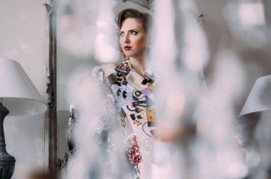 Kaliteli makyajlı, saç stili ve parlak renkli ceketli güzel sarışın kız aynanın yanında duruyor.