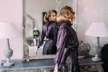 Kaliteli makyajlı, eski moda mor elbiseli güzel sarışın kız aynanın yanında duruyor ve ona bakıyor.