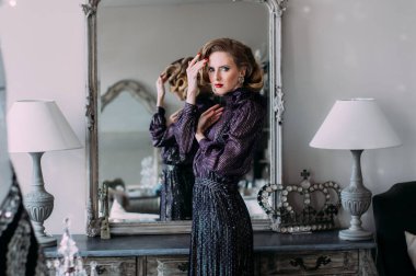 Klasik makyajlı, saç stili ve retro mor elbisesi olan güzel bir sarışın kız. Aynanın yanında duruyor ve kameraya bakıyor.