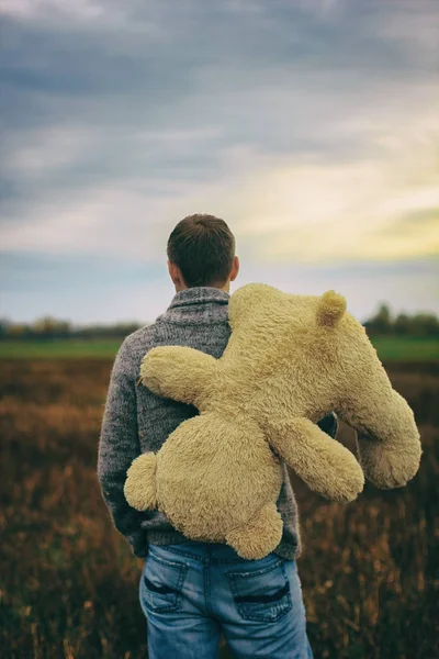 Um homem está segurando um urso de marionete atrás dele. Ele caminha pelo campo. Tristeza — Fotografia de Stock