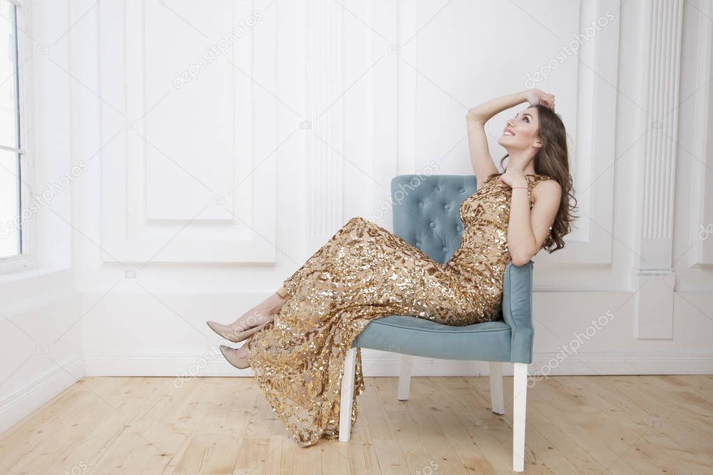 Young beauty woman in golden dress sitting in blue armchair, luxury apartments interior background 