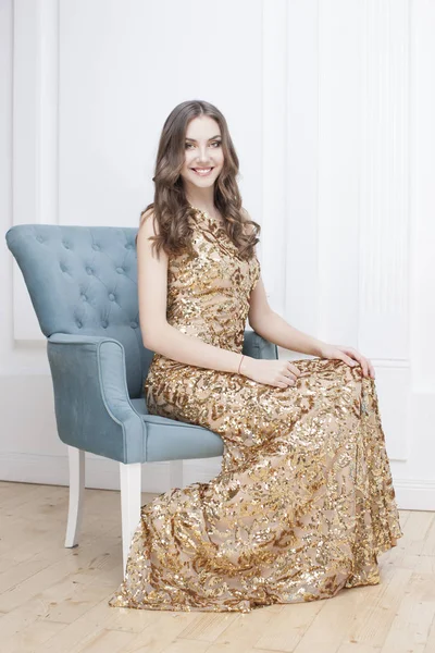 年轻的美女在金色礼服坐在蓝色扶手椅 豪华公寓内部背景 — 图库照片