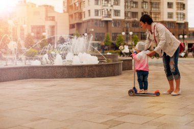 Küçük kız sürme scooter, annesi tarafından çeşme arka plan bulanık binalar ve şehir merkezine yürüyüş sırasında yardım ediliyor