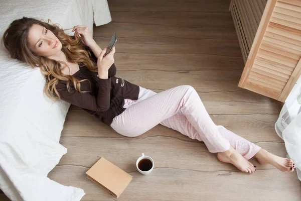 Pijama giymiş ve smartphone, yerde duran hoş kokulu kahve fincanı kullanarak pencere, yatak odasında katta oturan güzel kız